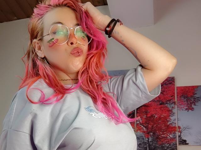 NatashaLau with pink and orange hair, puffy green-rimmed glasses, and a gray tee, sticking out her tongue, hand in hair, black bracelets, and a small nose piercing.