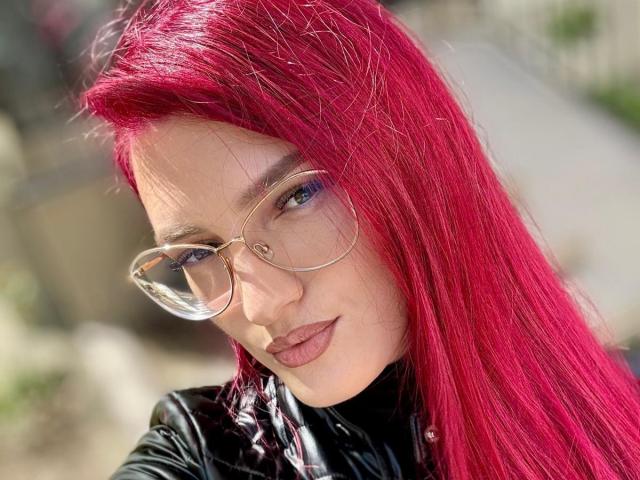 Rosalyn, avec ses cheveux rose vif, ses lunettes claires et son top en cuir noir, cligne de l'œil à la caméra, mettant en valeur son visage sexy et mignon, ainsi que son maquillage discret..