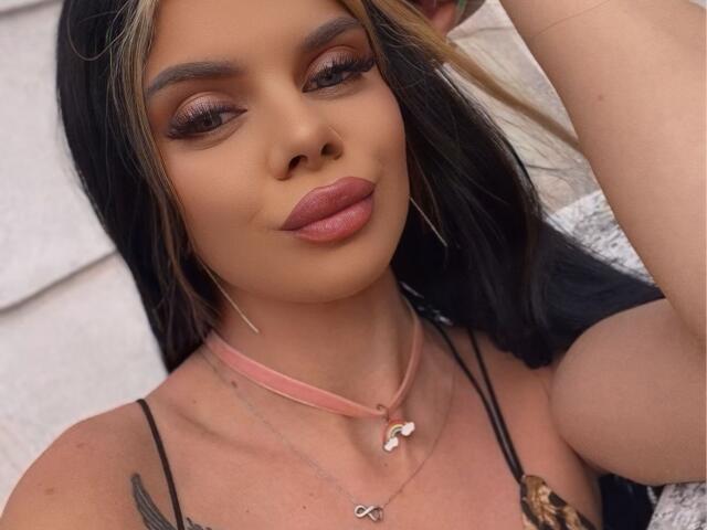 IvyRaven has tan skin, black and blonde hair, pink lipstick, and tattoos. She's wearing a pink choker with a heart and necklace. Boobs visible in black straps.