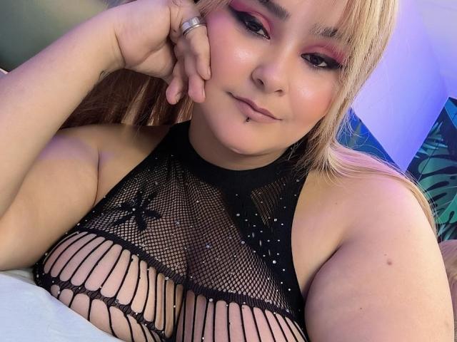 FlaviaSaenz with blonde hair, pink eyeshadow, and black mesh top, showing her large tits through the net, resting her chin on her hand, with a subtle smirk.