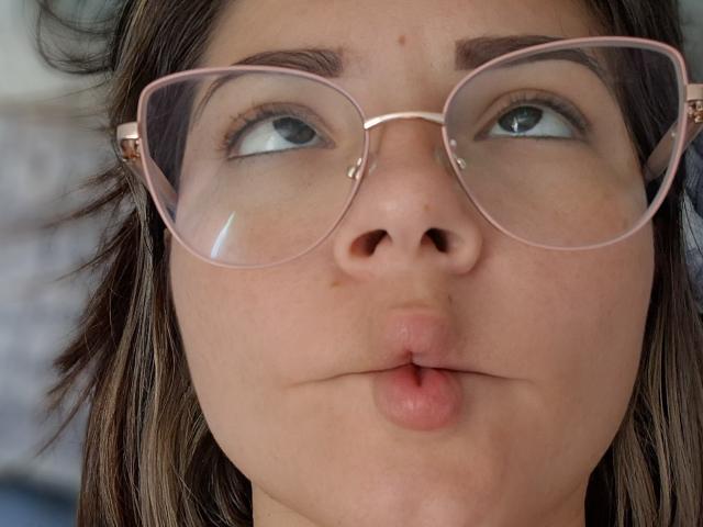 KatherineToro's pouty face with pink-rimmed glasses, brown eyes looking up, and slightly parted pink lips. She has light skin, dark brown hair, and small moles above her eyebrows.