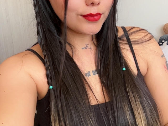 KimCoquine69 with long black hair in braids, red lipstick, black tank top, tattoos on neck and chest, visible arm tattoo, medium tits, light skin, no visible nose.