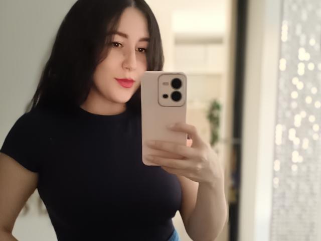 Maby Legrand, con lunghi capelli neri, rossetto rosa e una canottiera nera aderente, tiene in mano un iPhone rosa, sta scattando un selfie, mostrando il seno e un sorriso appena accennato..