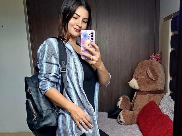 KatherineToro with dark hair, wearing a striped shirt, black tank, and backpack, holding phone with purple case, smiling at mirror, standing next to brown teddy bear and red pillow.