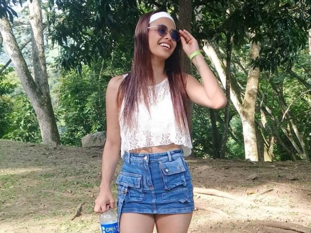 MaiteRosy with long brown hair, white lace crop top, blue denim skirt, white headband, sunglasses, smiling, holding a blue drink, standing in a forest, sunny, looking cute.
