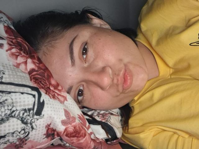 KatherineToro with puffy lips, brown eyes, and slight makeup, lying on a floral pillow, wearing a yellow hoodie, her face showing a neutral to slightly annoyed expression.