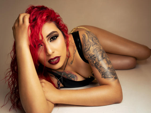 Eimy Callahan mit roten Haaren, schwarzem Oberteil, goldener Kette, tätowiertem Arm, liegend, Blick in die Kamera, dunkler Lippenstift, sexy, gebräunte Haut, Brüste sichtbar, Hand im Haar, verführerische Pose..