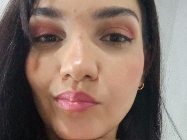 AngelyeRoss has pink eyeshadow, black eyeliner, and pink lipstick, with dark brown hair and a gold earring in her right ear, looking slightly pouty.
