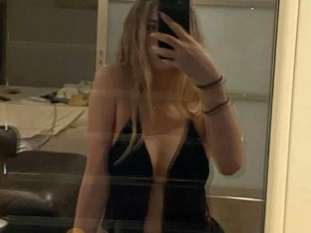 Mirror selfie of Lamour-hot with long blonde hair, black deep V-neck dress showing tits, holding phone, wearing bracelets, in dimly lit room.