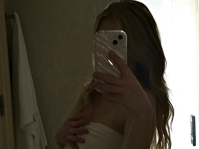 Lamour-hot with wavy blonde hair, holding a phone over her face, covering her tits with a white towel, in a dimly lit bathroom.