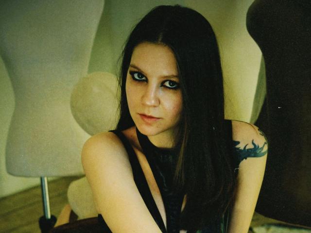 Celestai, met lang zwart haar, donkere eyeliner en een zwarte tatoeage op haar rechter schouder, draagt een zwarte top en kijkt verleidelijk in de camera..