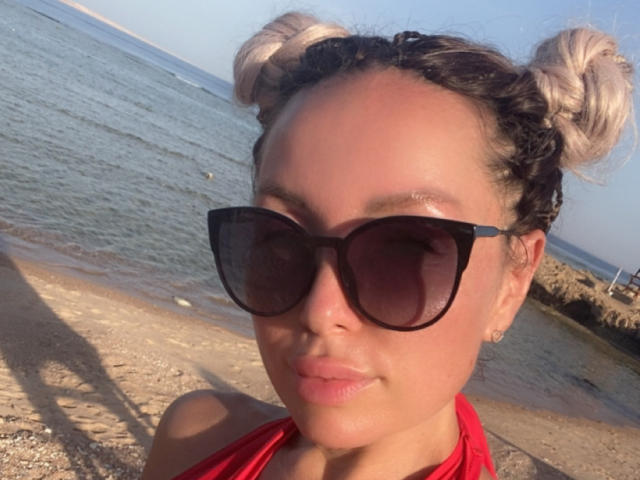 AvaGreenx with blonde double buns, black sunglasses, and red halter top, showing her cute face and subtle smile on a sunny beach. Her tits are not visible.