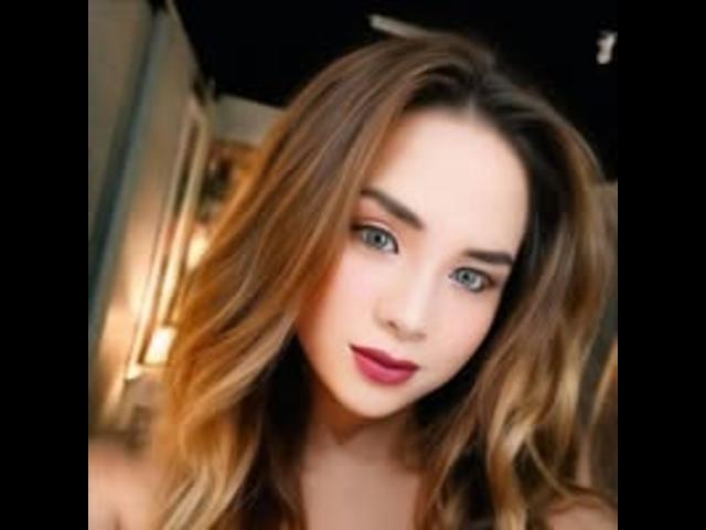 AlluringJhana has long, wavy brown hair, blue eyes, and pink lipstick, looking at the camera with a slight smile. Her tits are not visible, just her face.