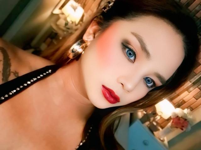 AlluringJhana has blue eyes, red lipstick, and a tattoo on her right shoulder. She's wearing a black halter top with silver studs. Her brown hair is partially up.