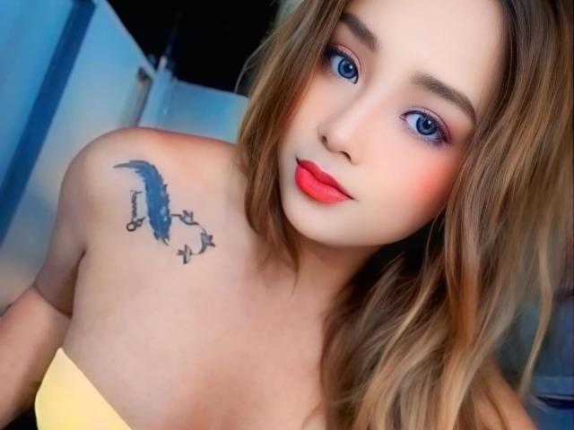 AlluringJhana has blue eyes, red lipstick, and a bird tattoo with small birds on her shoulder. She's wearing a yellow strapless top, with light brown, wavy hair.