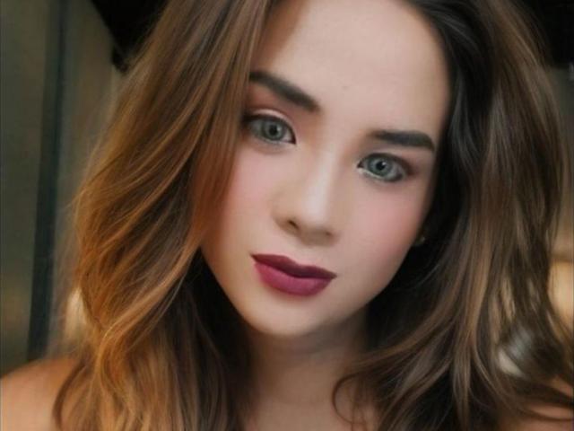 AlluringJhana has green eyes, wavy brown hair, and wears dark pink lipstick. Her eyebrows are thick and she's looking directly at the camera with a slight, confident smile.