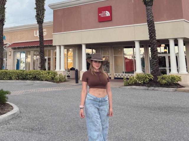 ArianaLu standing in front of a store, wearing a brown crop top, blue high-waisted jeans, and a gray hat, with her tits slightly visible and a pink watch.