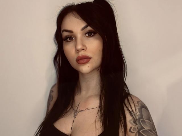 InkedFaye with long black hair, tattooed arms, and full pink lips, wearing a black top, looking directly at the camera with a neutral expression. She's a sexy, white woman.
