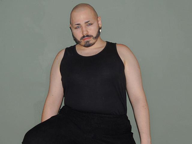 FabianNoLimits has a bald head, black tank top, black pants, and a beard with a small mustache. He's sitting with a neutral expression against a plain green background.