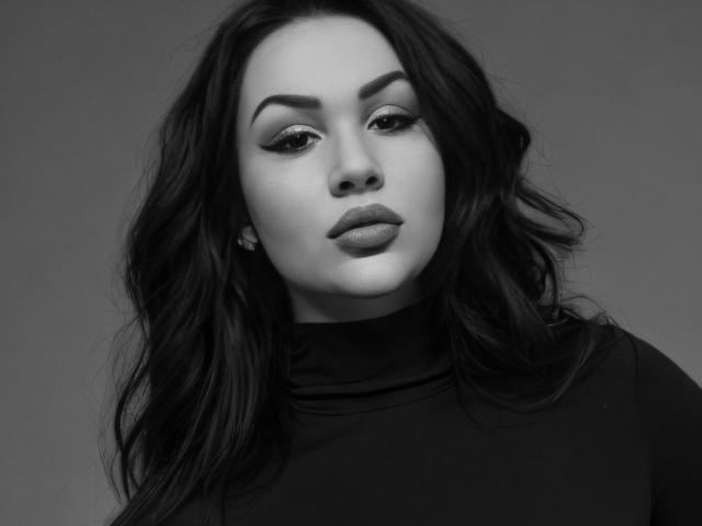 InkedFaye has full lips, dark wavy hair, and thick eyebrows, wearing a black turtleneck. Her eyes are half-open with subtle makeup. She's a sexy, black-and-white, grayscale photo.