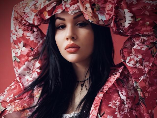 InkedFaye with long black hair, wearing a red floral shirt, hands in shirt, staring with full orange lips, choker necklace, and a sexy, confident expression.