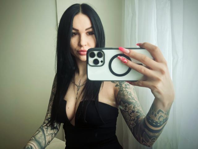InkedFaye with long black hair, tattoos on arms, pink nails, taking a selfie in a mirror, wearing a black dress, showing her boobs, holding a white phone.