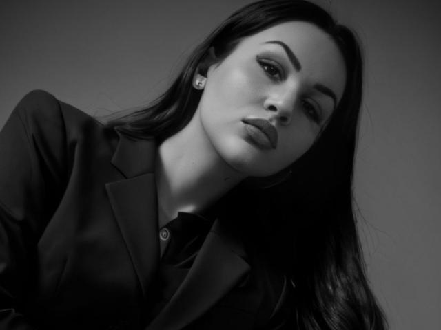 InkedFaye with dark, straight hair, wearing a black blazer, looking down with slightly parted, full lips and subtle makeup, has small, diamond earrings and a soft, gray background.