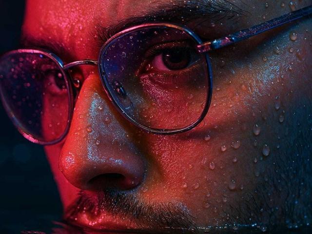 OrfeoJames's sweaty face, red and blue lighting, wearing round glasses with droplets, intense gaze, slight stubble, dark background, focus on nose and glasses, serious expression.