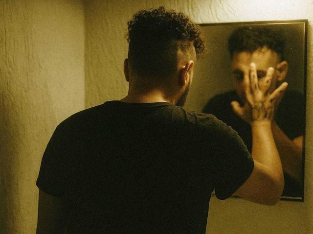 OrfeoJames with curly hair and black t-shirt, staring at mirror, hands over face, reflection shows worried expression, dimly lit, textured wall in background.