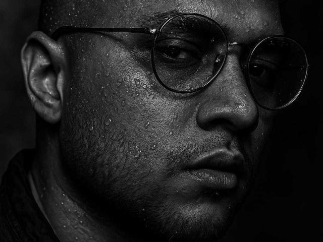 OrfeoJames's sweaty, dark-skinned face with droplets, wearing round glasses, has a serious expression and a slight beard, in a black-and-white, high-contrast photo.