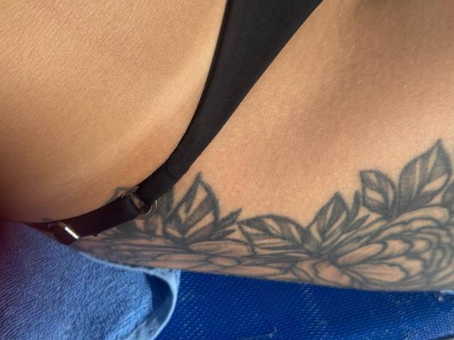 Tsukerbergs schwarzer String ist zur Seite gezogen, wodurch ein detailliertes Tattoo von blattartigen Blumen auf ihrem gebräunten, kurvigen Hintern sichtbar wird. Der Metallring des Strings ist zu sehen..