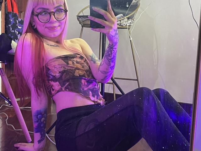 KallySpice with pink hair, glasses, tattoos, strapless top with a skull print, black sparkly pants, sitting, taking a mirror selfie, smiling, showing her ass, and holding a phone.