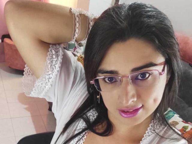 KhalifaGold with glasses, purple lipstick, and white lace top, raising arm to show off tits, dark hair, and brown skin, sitting in a room.