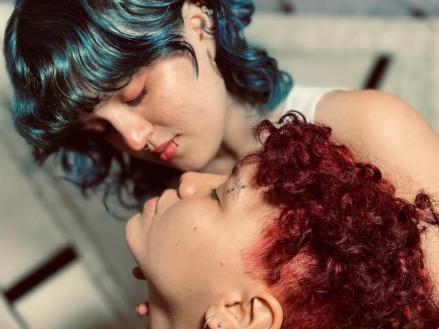 JessAndJade with blue and black curly hair kissing the red curly-haired JessAndJade's nose, both with nose piercings, in an intimate, close-up shot.