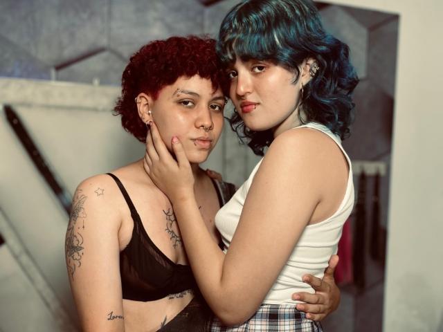 JessAndJade with curly red hair and black bra, holding blue-haired JessAndJade's face, wearing white tank top and plaid skirt, both showing tattoos, standing closely, intimate.