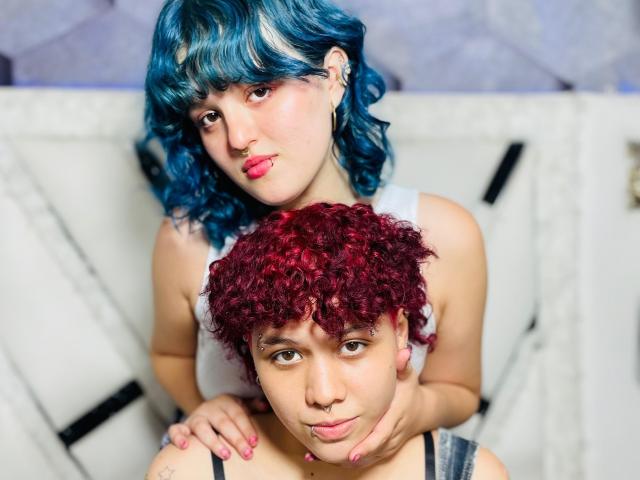 JessAndJade with blue curly hair and nose ring, holding the red curly-haired JessAndJade's chin, both wearing tank tops, sitting on white couch. JessAndJade's pink lipstick stands out.