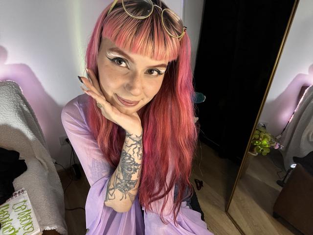 KallySpice with pink and yellow hair, eyeshadow, tattooed arm, purple shirt, glasses on head, smiling, hand on cheek, sitting, wood floor, mirror, pink lights.