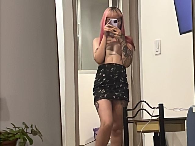 KallySpice with pink hair, topless, showing small tits, wearing a black floral skirt, taking a mirror selfie with a phone, tattoo on right arm.