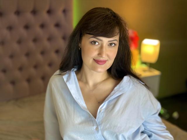 NathalyStars with medium tits, wearing an unbuttoned light blue shirt, sitting on a bed, has long dark brown hair, slight smile, and green eyes, in a softly lit room.