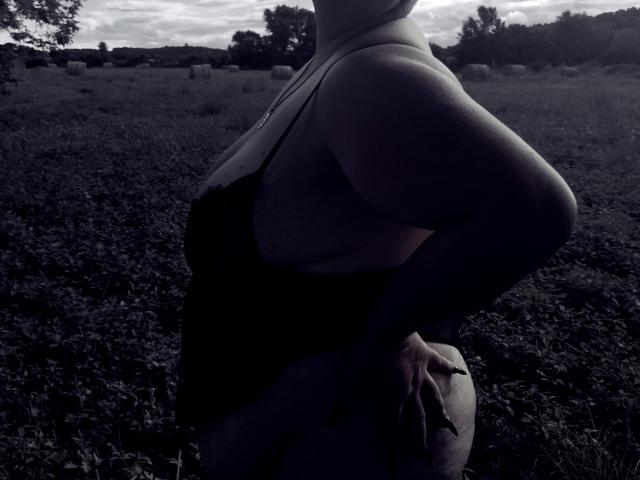 Das Profil von MmeCauchemar, ein dunkles Tanktop, große Brüste, Hände an den Hüften, stehend in einem schattigen, grasbewachsenen Feld, bewölkter Himmel, Bäume in der Ferne, monochrom, leicht überbelichtet..