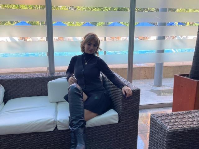 Marianne Bowe, mit kurzen, braunen Haaren, trägt einen schwarzen Rollkragenpullover und einen schwarzen Rock. Sie sitzt in einem Korbstuhl mit weißen Kissen, schwarzen Stiefeln und einer goldenen Halskette..