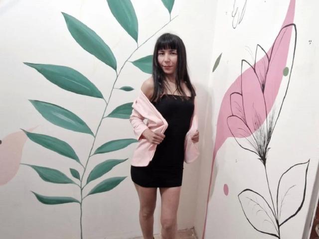 MilagrosPop, met lang zwart haar, draagt een strak zwart minirokje en een roze jas, en staat in een kamer met groene en roze bloemenmotieven op de muur. Haar achterkant is zichtbaar..
