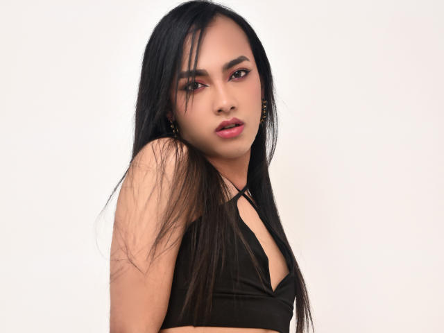 YelianaSantana with long black hair, wearing a black halter top, exposing small tits, has a serious expression, and wears gold earrings, against a plain white background.