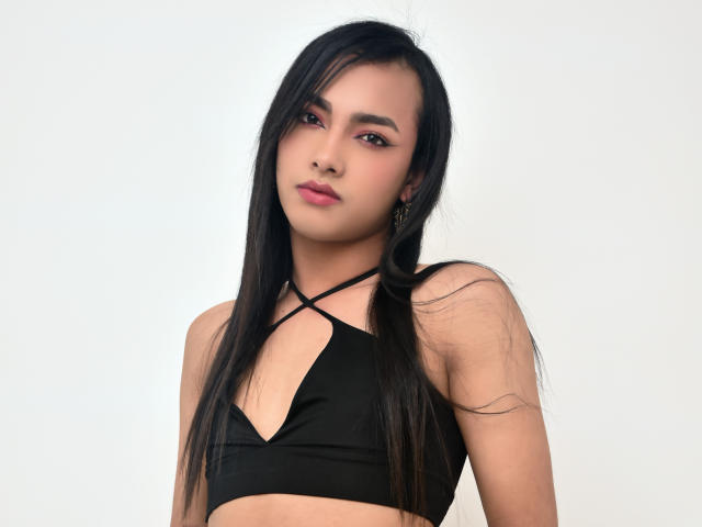 YelianaSantana with long black hair, pink eyeliner, and black strappy top, showing small tits, looking at camera with slight tilt, white background.