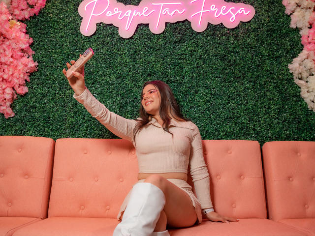EmmaMack with long brown hair, tan crop top, white knee-high boots, taking a selfie with a pink dildo, sitting on a pink couch, smiling.