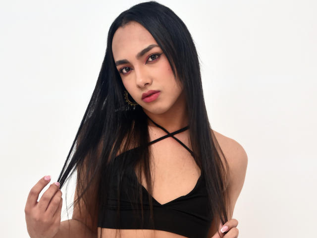 YelianaSantana with long black hair, wearing a black crop top with crisscross straps, holding her hair, looking at the camera, wearing pink lipstick and gold earrings.