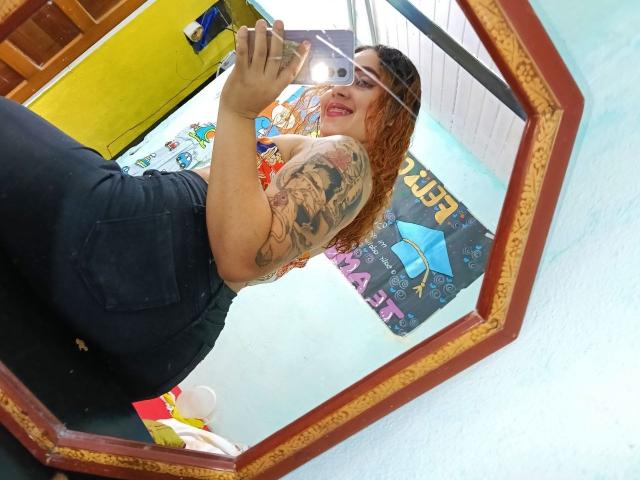 MerydaBraveI with tattoos, red lipstick, black high-waisted pants, and a phone in a hexagonal mirror, showing her butt and smiling. Her ass is prominent.