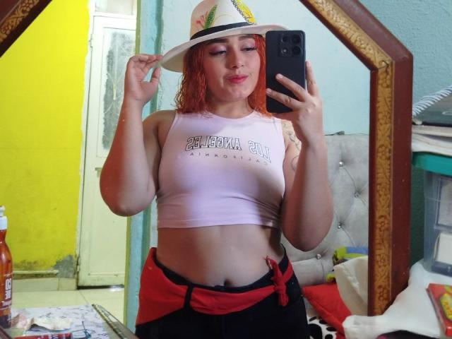 MerydaBraveI with red curly hair, white hat, pink crop top, showing her tits, holding phone, mirror selfie, red and black skirt, room with yellow wall.