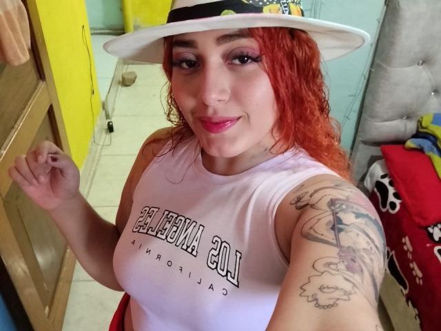 MerydaBraveI with red curly hair, white hat, pink lipstick, white sleeveless top, tattooed arm, showing her tits, standing in a room with yellow and blue walls.