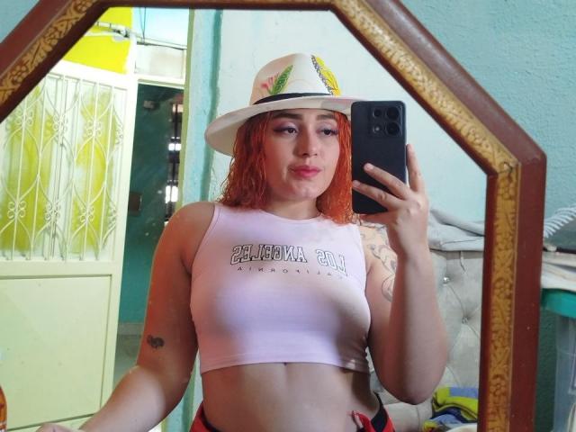MerydaBraveI with red hair, white hat, and pink crop top, showing her tits in a mirror selfie, holding a black phone, with a tattoo on her left arm.
